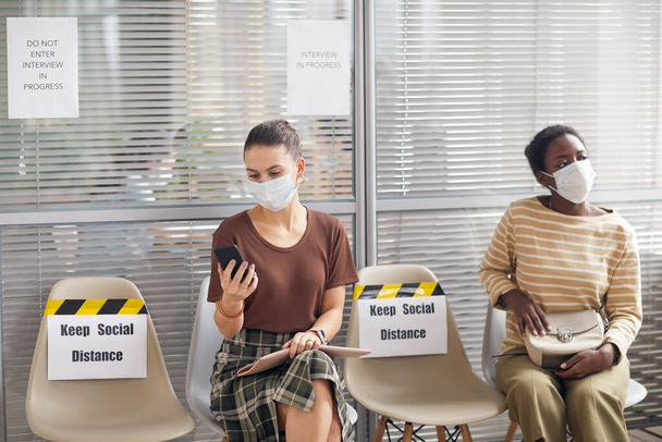 Портрет двух женщин в масках, стоящих в очереди в офисе с табличкой "Держите социальную дистанцию" во время эпидемии, скопируйте пространство - Фото, изображение