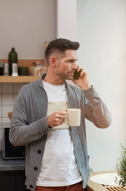 Vertical waist up portrait of handsome mature man speaking by smartphone and enjoying cup of coffee in morning while standing by window in kitchen - 写真・画像