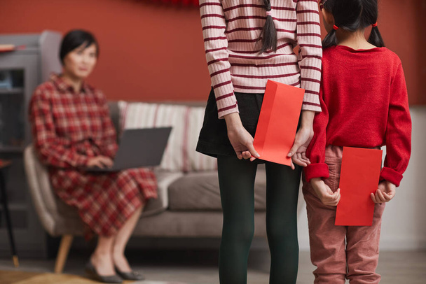 Dos irreconocibles chicas asiáticas de pie en la sala de estar frente a su madre escondiendo regalos sobre rojo para ella a sus espaldas - Foto, imagen