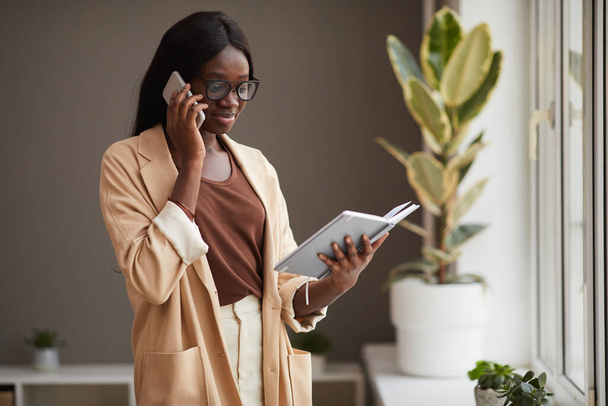 Талия портрет успешной афроамериканской предпринимательницы, говорящей по телефону в офисе и держащей в руках открытую тетрадь при планировании деловых встреч, копирования пространства - Фото, изображение