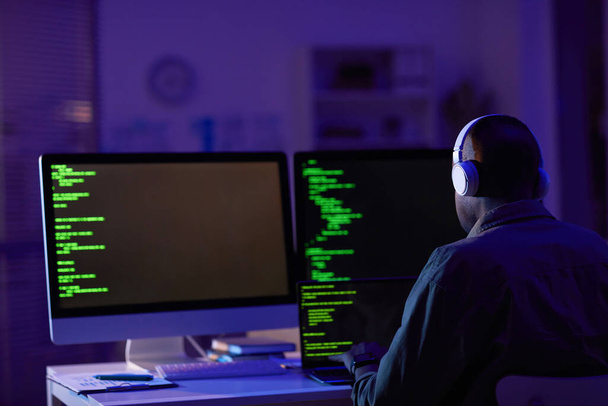 Снимок с заднего вида профессионального разработчика программного обеспечения в наушниках, сидящих в офисе в одиночестве и работающих на компьютерах, создающих новый код - Фото, изображение