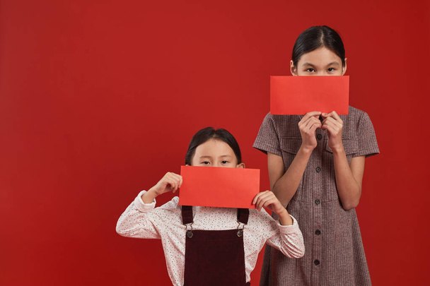 Studioporträt zweier asiatischer Mädchen, die vor rotem Wandhintergrund stehen und Gesichter hinter roten Umschlägen verstecken, Kopierraum - Foto, Bild