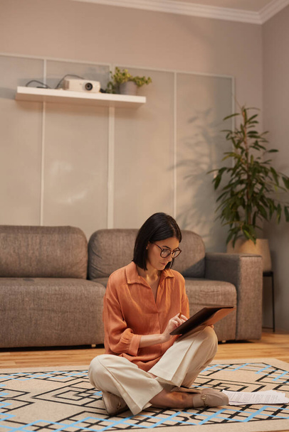 Full length portrait of contemporary woman working from home while sitting cross legged on floor and using digital tablet - Photo, image