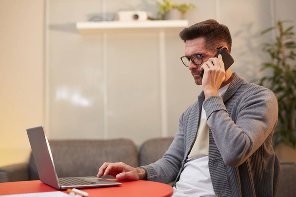 Widok z boku portret przystojnego dojrzałego mężczyzny pracującego z laptopem i mówiącego przez telefon siedząc przy czerwonym stole w minimalnym pomieszczeniu biurowym, przestrzeń do kopiowania - Zdjęcie, obraz