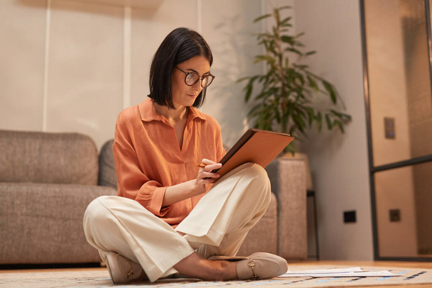 Full length portrait of contemporary woman working from home while sitting cross legged on floor and using digital tablet, copy space - Foto, Bild