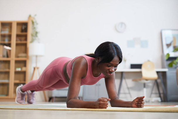 Teljes hosszúságú portré fiatal afro-amerikai nő csinál deszka gyakorlat, miközben élvezi az edzés otthon, másolás hely felett - Fotó, kép
