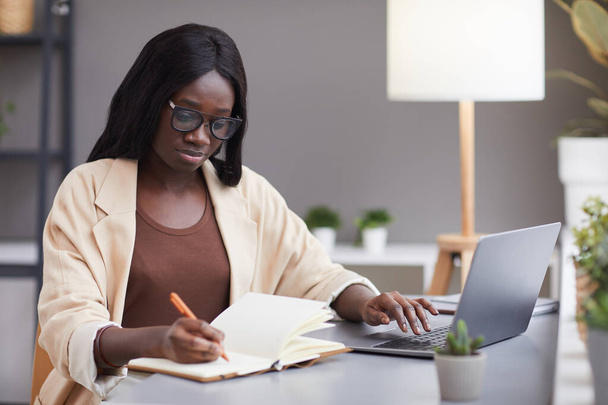 Porträt der schönen afroamerikanischen Geschäftsfrau, die Laptop benutzt und Notizen macht, während sie erfolgreiche Geschäfte führt oder studiert, Kopierraum - Foto, Bild
