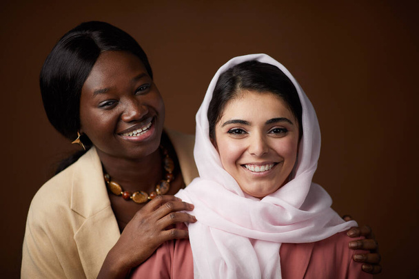 Minimální portrét dvou etnických mladých žen usmívajících se před kamerou, pózujících na hnědém pozadí ve studiu a oslavujících dědictví a osobní identitu - Fotografie, Obrázek