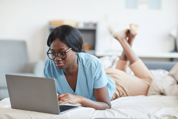 Retrato de una joven afroamericana que usa una computadora portátil mientras está acostada en la cama y disfruta del fin de semana en casa con comodidad, espacio para copiar - Foto, imagen