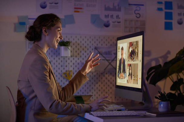 Πλάγια άποψη φωτογραφία πορτρέτο της χαρούμενης νεαρής γυναίκας που περνά το βράδυ στο γραφείο λαμβάνοντας μέρος σε online συνάντηση με τους συναδέλφους - Φωτογραφία, εικόνα