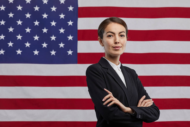 Талия портрет уверенной женщины-политика, смотрящей в камеру, стоя с оружием, скрещенным на фоне флага США, копировальное пространство - Фото, изображение