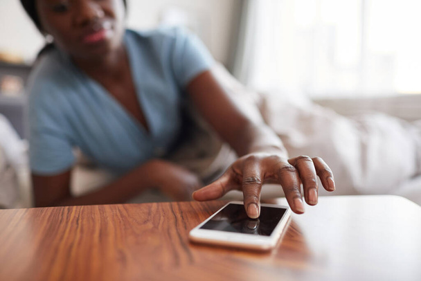 Close-up van onherkenbare Afrikaans-Amerikaanse vrouw tikken smartphone op nachtkastje terwijl het uitschakelen van wekker of het controleren van berichten in de ochtend, kopieer ruimte - Foto, afbeelding