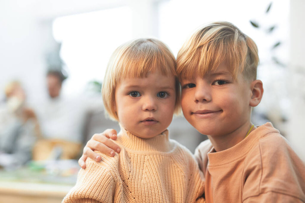 Portrét roztomilé blondýny chlapec objímající sestřičku a při pohledu na fotoaparát, zatímco si užívají čas s rodinou doma, kopírovat prostor - Fotografie, Obrázek