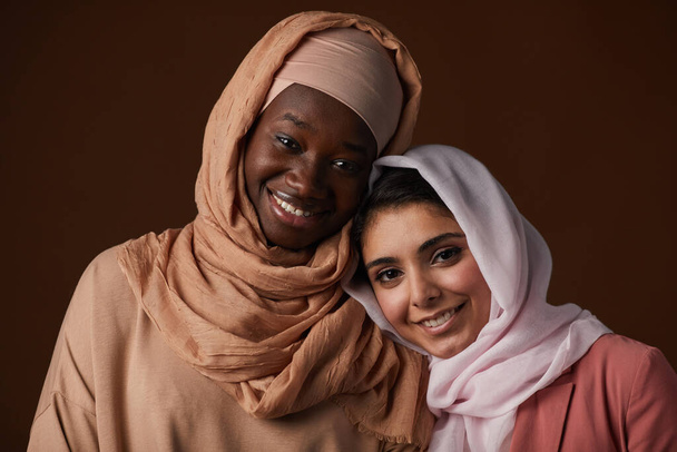 Retrato de duas jovens étnicas usando lenços de cabeça e sorrindo para a câmera enquanto posando contra fundo marrom no estúdio, celebrando herança e identidade pessoal - Foto, Imagem