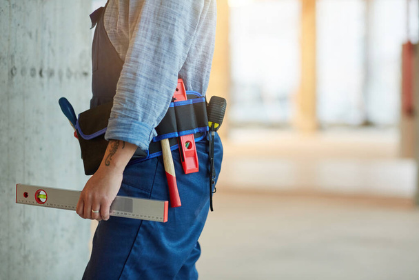 Cropped side view of modern female worker holding level and wearing toolbelt while working on construction site, copy space - 写真・画像