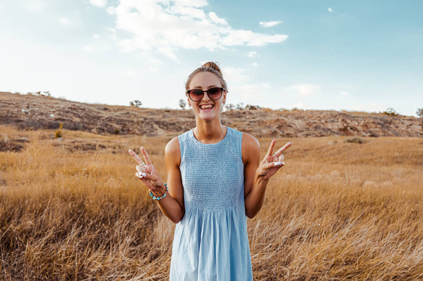 χαρούμενη όμορφη νεαρή γυναίκα με καλοκαιρινό φόρεμα και γυαλιά ηλίου που δείχνουν σύμβολα ειρήνης και ποζάρουν στο λιβάδι  - Φωτογραφία, εικόνα