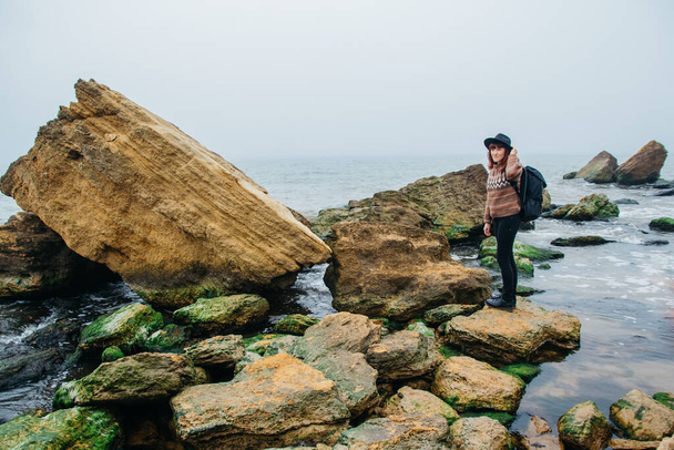 Eine junge Touristin mit Hut und Rucksack steht auf einem Felsen vor einem schönen Meer, blickt aufs Meer, auf den Horizont. Platz für Text oder Werbung. - Foto, Bild