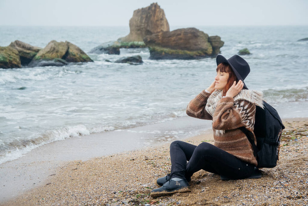 Kaunis nainen hattu ja huivi reppu istuu rannikolla taustaa vasten kiviä vastaan kaunis meri. Teksti- tai mainospaikka. - Valokuva, kuva