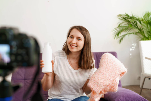 pretty female blogger sits on the couch in front of the camera and shows subscribers both a white bottle of fabric softener and a soft sweater washed with the help of the product. - Zdjęcie, obraz