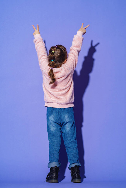 stylish little girl in fluffy sweatshirts and jeans stands with her back turned to full height on a purple background. Shows the victory sign in. Victory symbol. Emotional image. - Foto, Imagem