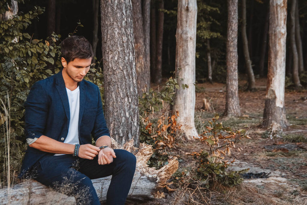 森の中で屋外でポーズをとっているハンサムな男が、チェックされたジャケットを着て丸太の上に座っている。テキストまたは広告の場所. - 写真・画像