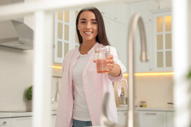 Young woman holding glass of pure water in kitchen, view through window - Фото, зображення