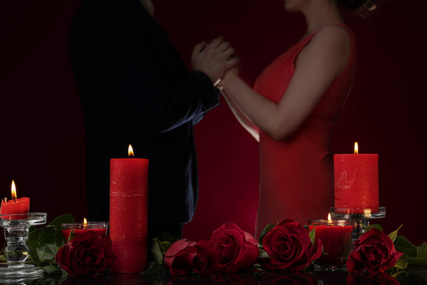 Ένας άντρας με σακάκι και μια γυναίκα με κόκκινο φόρεμα κρατιούνται χέρι χέρι. Κεριά και πολυτελή τριαντάφυλλα. Crop view of a romantic evening, Ημέρα του Αγίου Βαλεντίνου 14 Φεβρουαρίου, αρραβώνας, επέτειος γάμου, προοίμιο για τη νύχτα της αγάπης. - Φωτογραφία, εικόνα