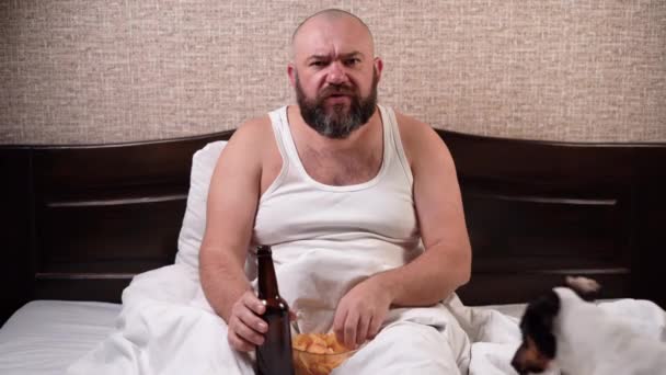 un hombre barbudo de 40 años se sienta en la cama en casa bebiendo cerveza come patatas fritas. mira las noticias con sorpresa. - Imágenes, Vídeo