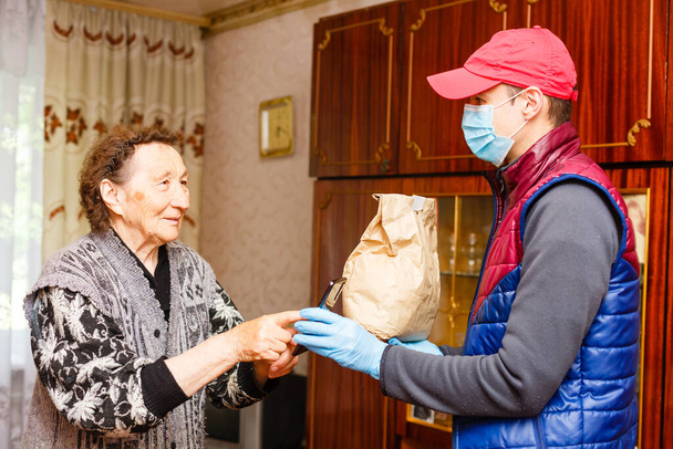 Une femme âgée reste à la maison. Livraison de nourriture dans un masque médical aux personnes âgées. - Photo, image