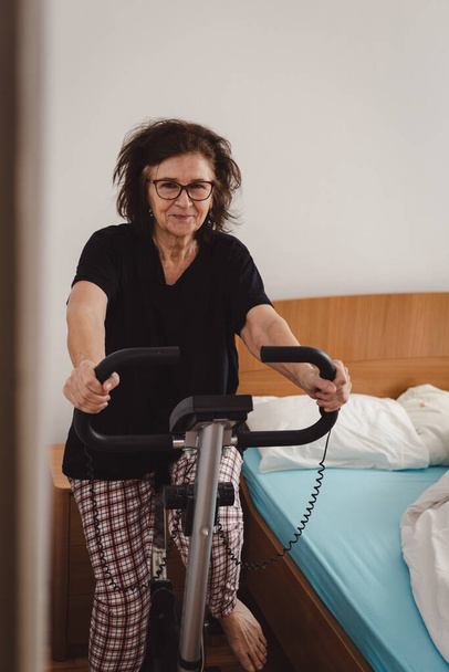 Par la porte de la chambre à coucher vue sur belle et souriante femme âgée au milieu des années 70 exercice sur vélo stationnaire à la maison. Femme mature active.  - Photo, image