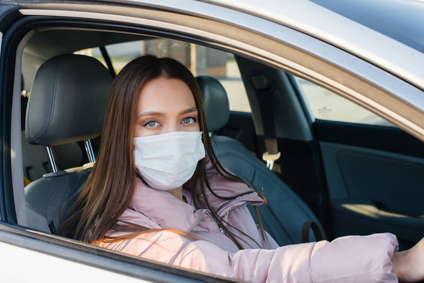 A young girl sits behind the wheel in the car in the mask during the global pandemic and coronavirus. Quarantine - Photo, Image