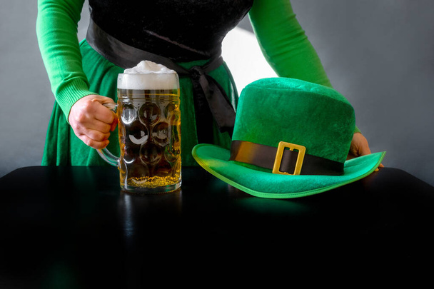 Main de femme tenant un verre de bière près du chapeau de Saint-Patrick d'un lutin sur le fond noir - Photo, image