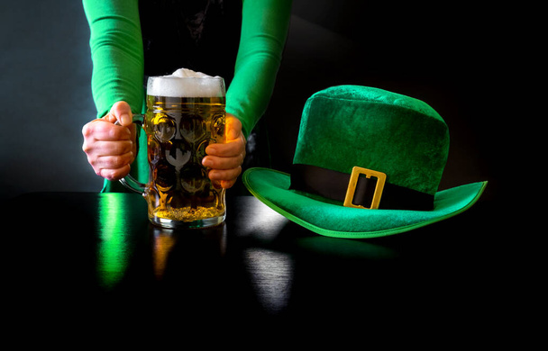 Die Hand einer Frau hält ein Glas Bier in der Nähe von St. Patrick 's Day Hut eines Kobolds auf schwarzem Hintergrund - Foto, Bild