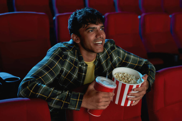 Portrait of excited young guy smiling, holding a drink and popcorn basket while watching movie alone in empty theater auditorium - Zdjęcie, obraz