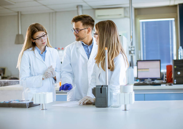 Skupina mladých vědců v ochranných pracovních oděvech stojících v laboratoři a analyzujících tekuté vzorky na iontových chromatografických zařízeních - Fotografie, Obrázek