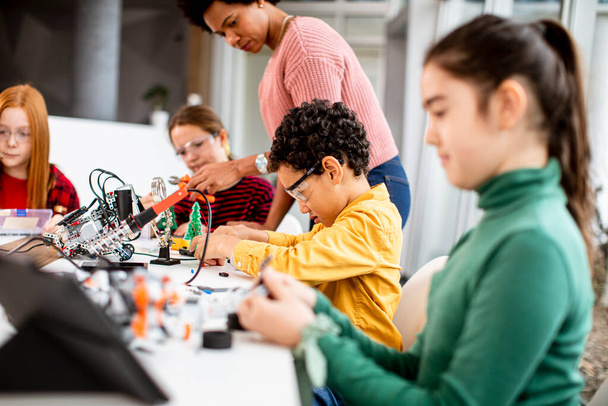 Улыбающаяся афроамериканка-преподаватель естественных наук с группой детей, программирующих электрические игрушки и роботов в классе робототехники - Фото, изображение