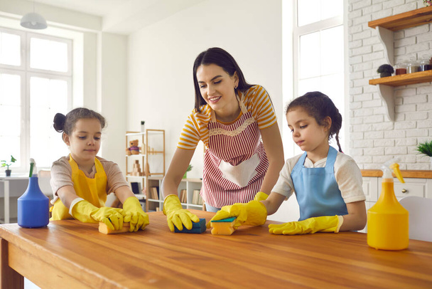 Happy little daughters helping their mom wash the table with sponges and detergent - Foto, imagen