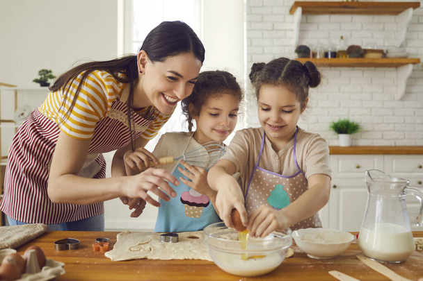 Happy young mother and cute helpful little daughters making dough for cookies together - Fotoğraf, Görsel