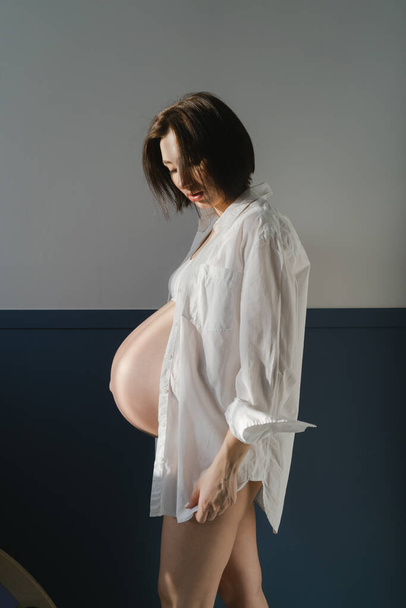 Νεαρή έγκυος κοπέλα με λευκά εσώρουχα και λευκό πουκάμισο κοντά σε λευκό τοίχο - Φωτογραφία, εικόνα