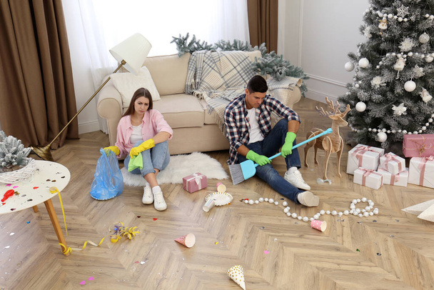 Tired couple sitting in messy room while cleaning after New Year party - Photo, image