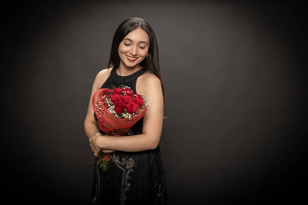 Όμορφη νεαρή μελαχρινή γυναίκα σε μαύρο φόρεμα ευτυχισμένη με ένα μπουκέτο κόκκινα τριαντάφυλλα. Ημέρα του Αγίου Βαλεντίνου. - Φωτογραφία, εικόνα