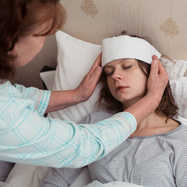 Νεαρή γυναίκα με υψηλή θερμοκρασία στο κρεβάτι με κλειστά μάτια, η μητέρα της κάνει συμπίεση σε χαμηλή θερμοκρασία. - Φωτογραφία, εικόνα