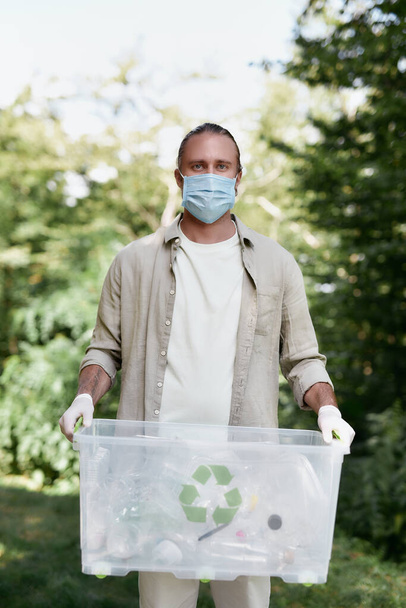 Recycling and COVID19. Young male eco activist wearing protective face mask with recycle bin looking at camera while collecting plastic waste in the forest during coronavirus pandemic - Фото, изображение