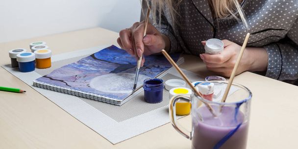 Un gros plan des mains féminines sur une image peinte tenant un pinceau et un couvercle pour aquarelles sur lequel les couleurs sont mélangées à côté de la gomme et un simple crayon. Un hobby de l'art. - Photo, image