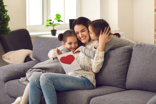 Happy young mother sitting on sofa and hugging two small beautiful kissing her daughters - 写真・画像