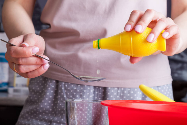 Female hands hold spoon and bottles of ready-made lemon juice in and pour it into the kneading dough from cottage cheese and flour in a red bowl against the background of eggs in a yellow substrate.  - Fotoğraf, Görsel