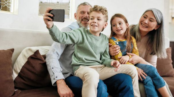 Catching bright moments. Happy grandparent with two cute kids grandchildren making a selfie on smartphone, relaxing together on sofa in the living room - 写真・画像