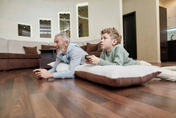 Online competiotion. Side view of a grandfather with grandson lying on pillows on the floor and playing video games, spending time together at home - Foto, imagen
