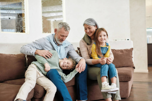 Portrait of lovely happy family, grandparents and grandchildren having fun and relaxing while sitting on sofa in the living room - Фото, изображение