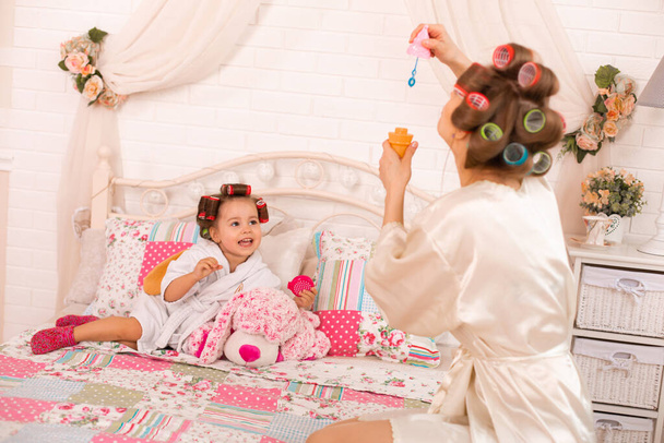 A charming little girl with her mother in hair curlers play with soap balls. Women's day. The girl has fun with her mom. - Photo, Image